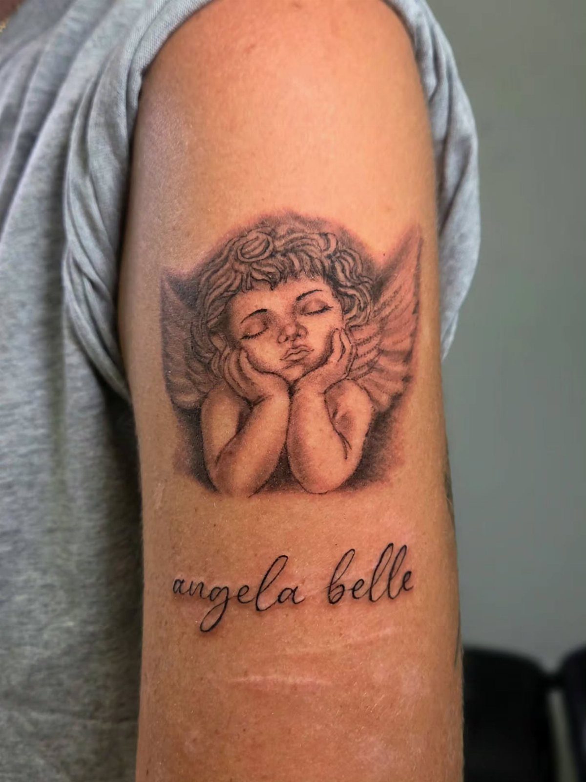 Cherub angel tattoo with wings and cursive text on a person's upper arm