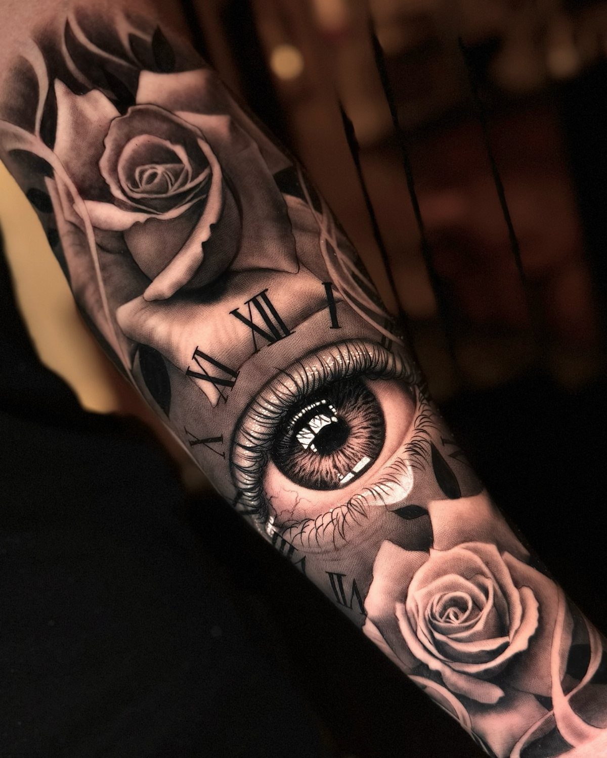 Black and grey tattoo on an arm featuring roses, an eye, and Roman numerals.