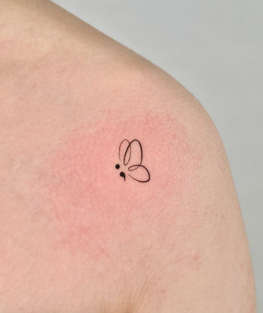 Butterfly semicolon tattoo in black ink on a person's shoulder with red skin.