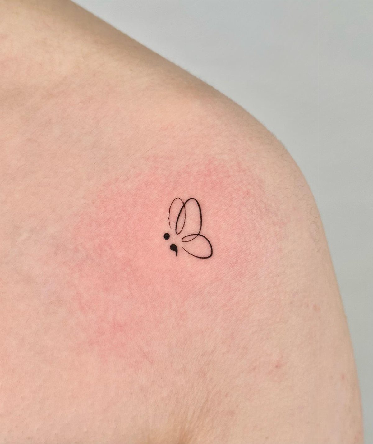 Butterfly semicolon tattoo in black ink on a person's shoulder with red skin.