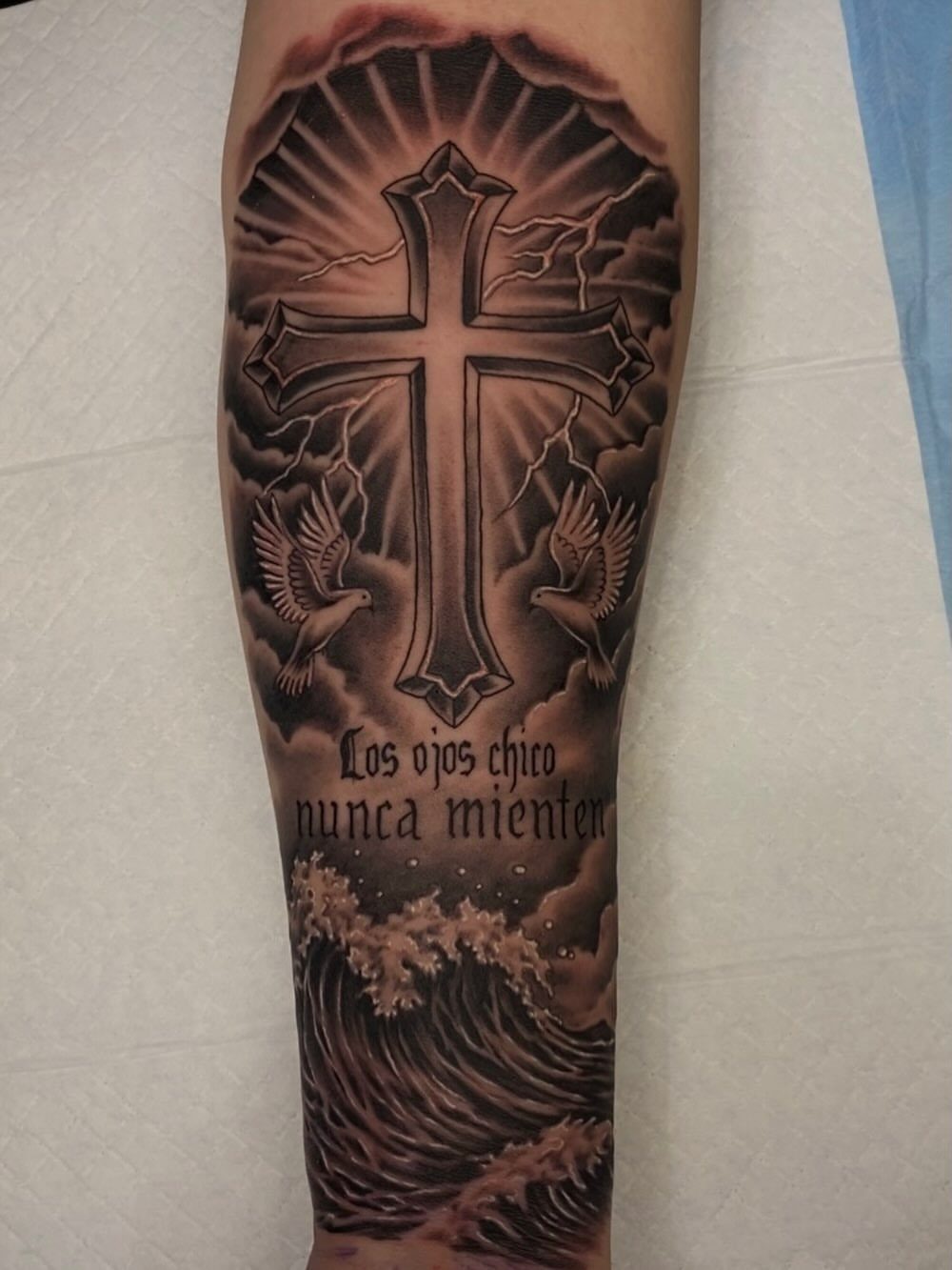 Cross forearm tattoo with doves, Spanish text, and ocean waves in black ink