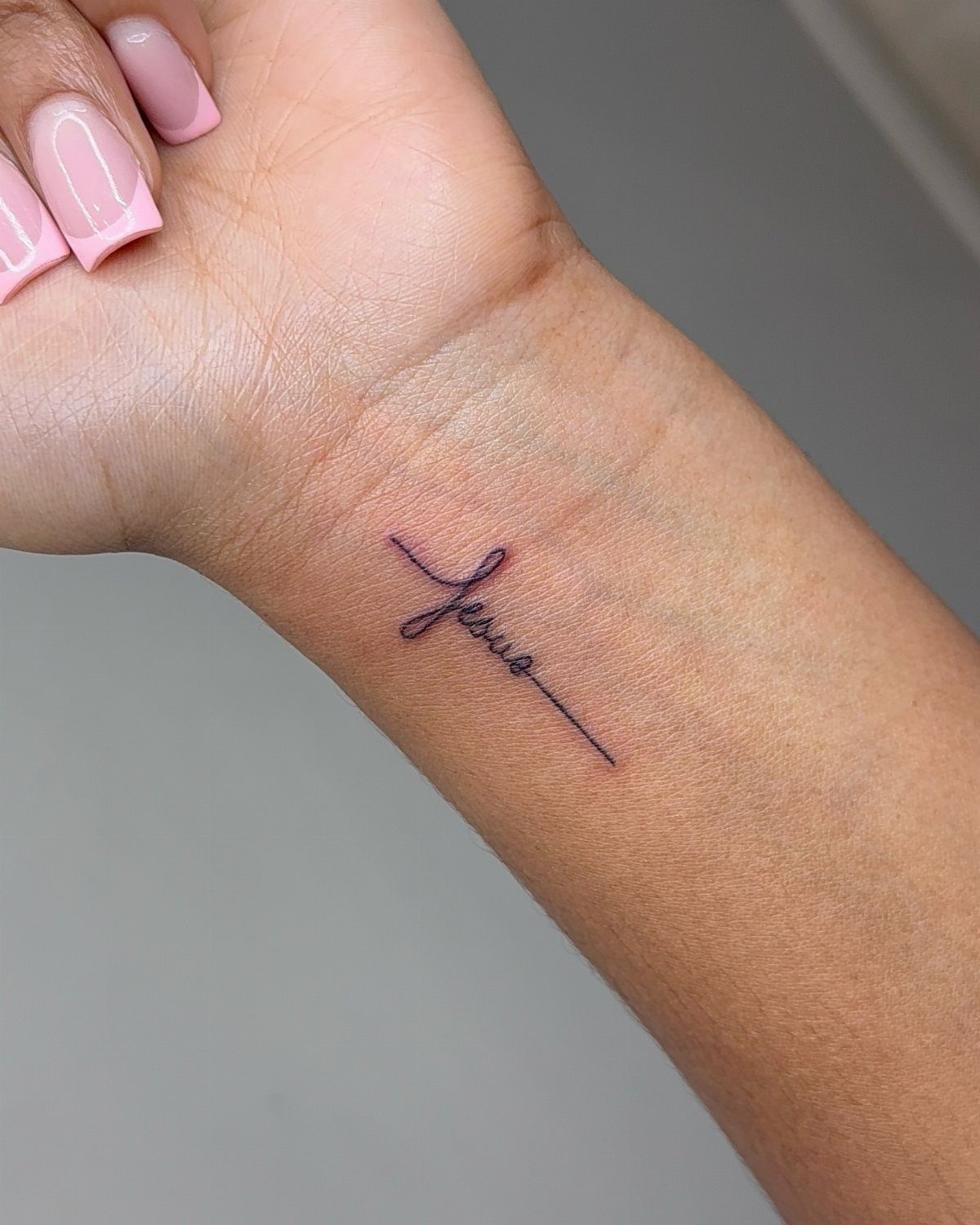 Small cross tattoo with the word Jesus in cursive on a wrist