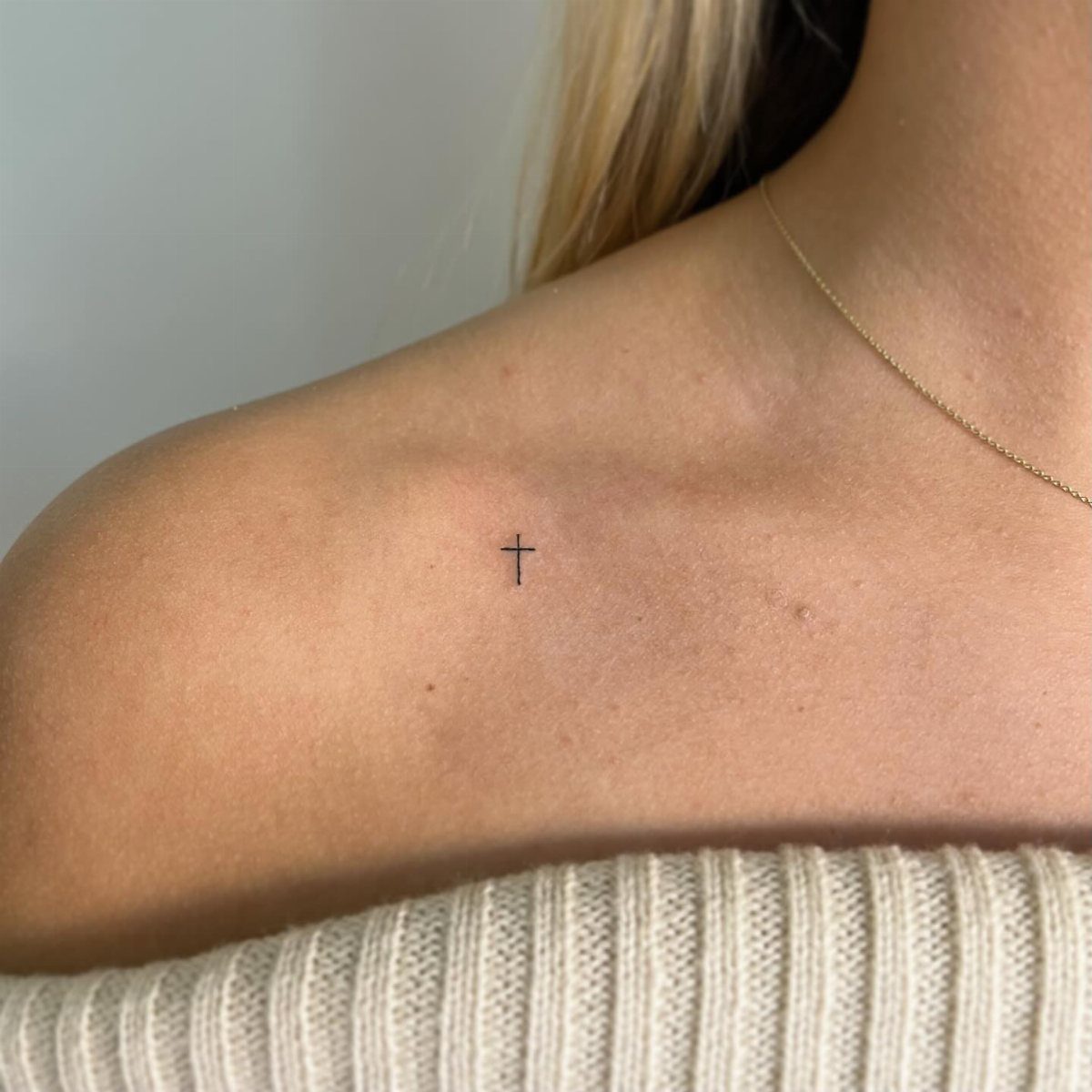 Small black cross tattoo on a woman's shoulder with a gold necklace.