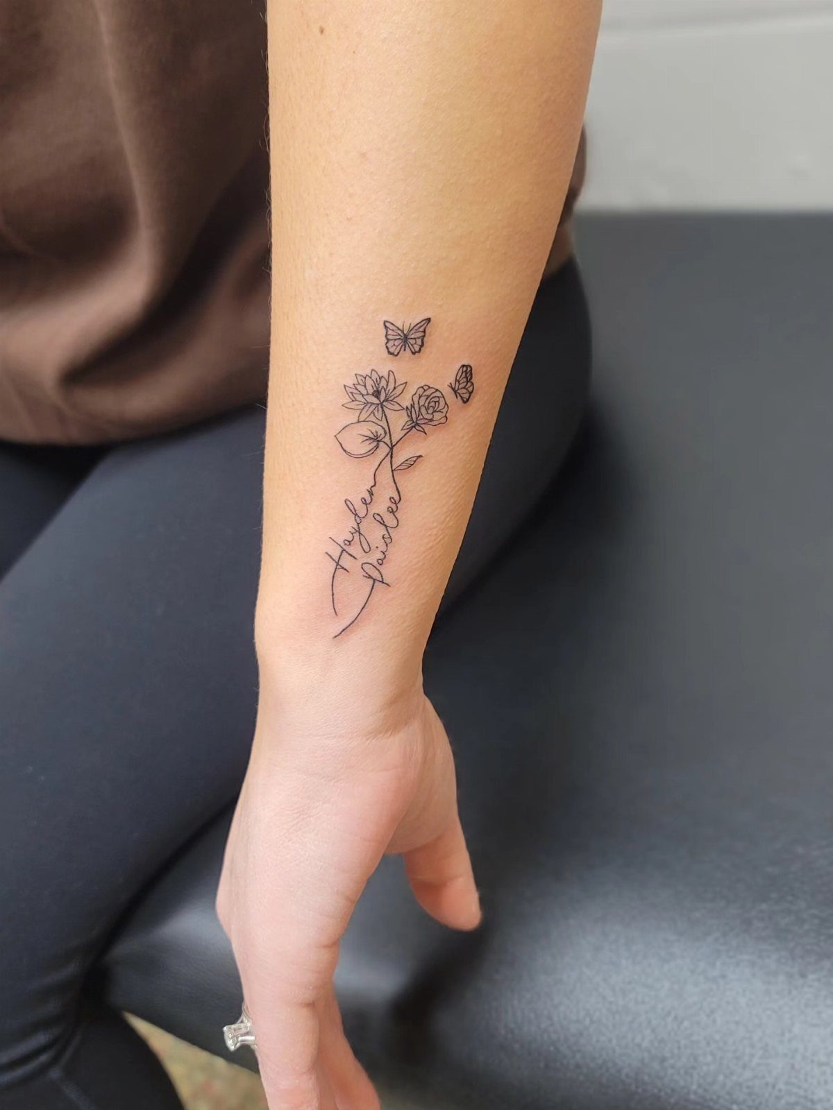 Forearm tattoo featuring delicate black ink flowers, butterflies, and cursive names on stem.