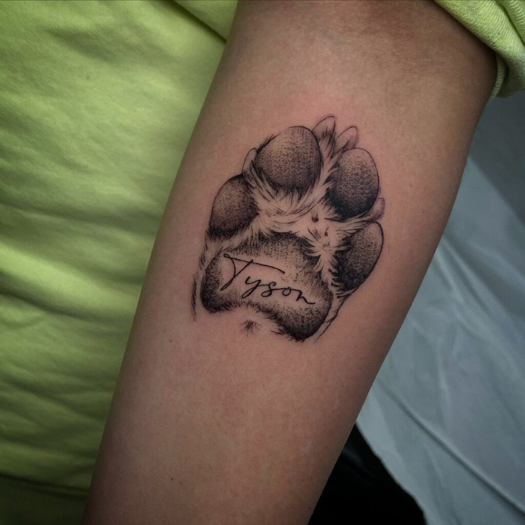 Dog paw print tattoo on a forearm with the name Tyson written inside.