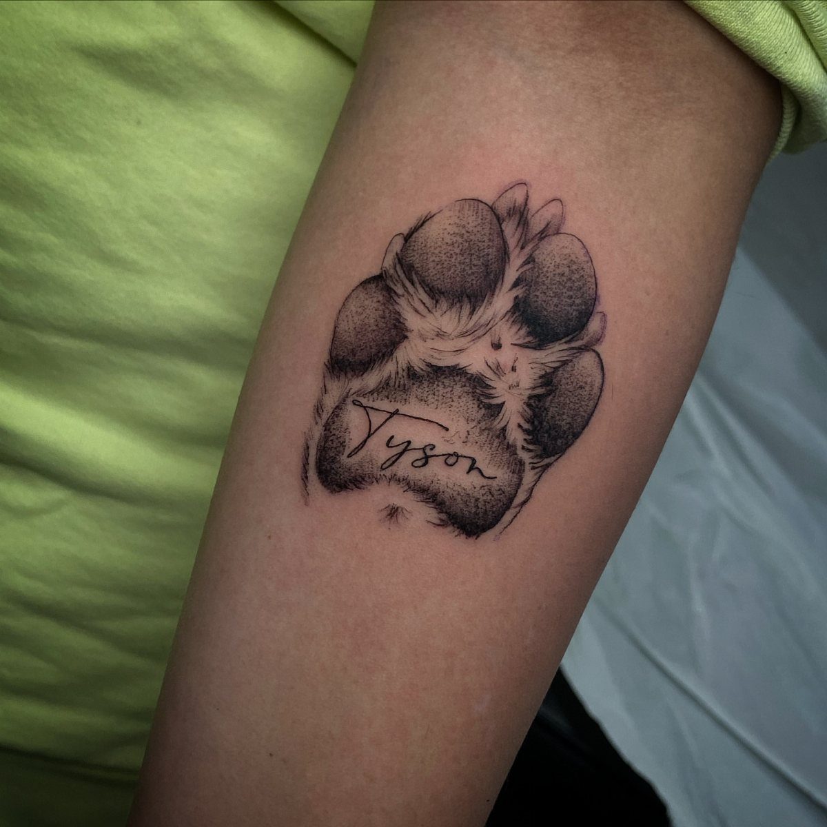 Dog paw print tattoo on a forearm with the name Tyson written inside.