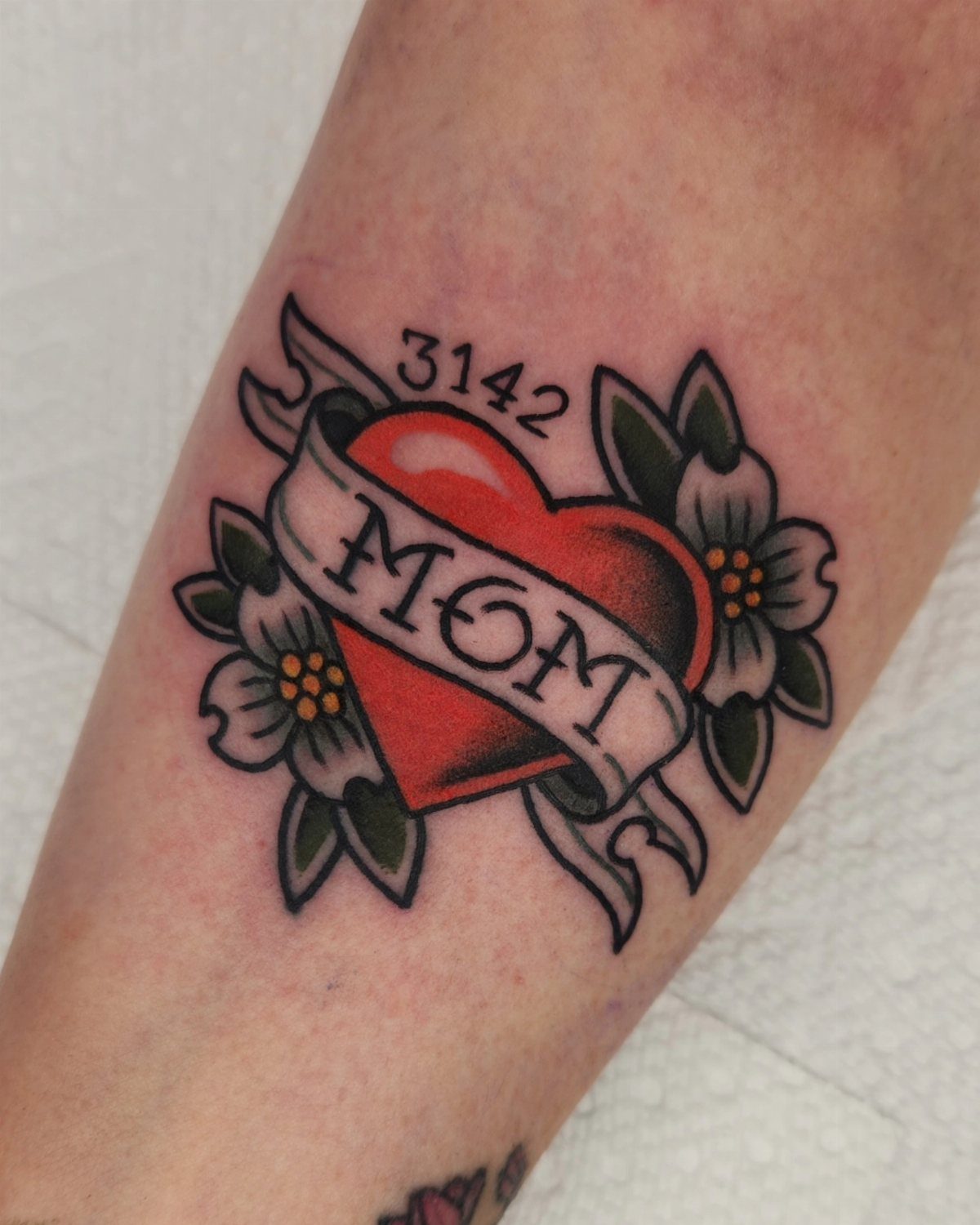 Traditional red heart tattoo with MOM banner and flowers on a forearm