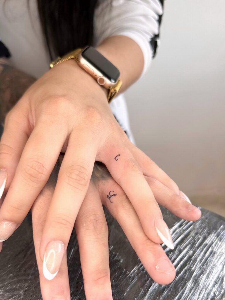 Two hands with small black initial tattoos on the sides of their ring fingers.