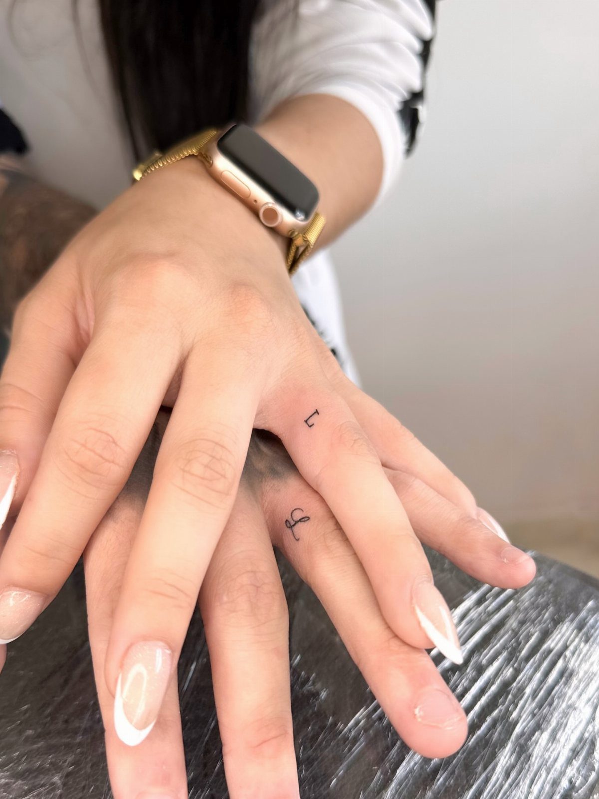 Two hands with small black initial tattoos on the sides of their ring fingers.