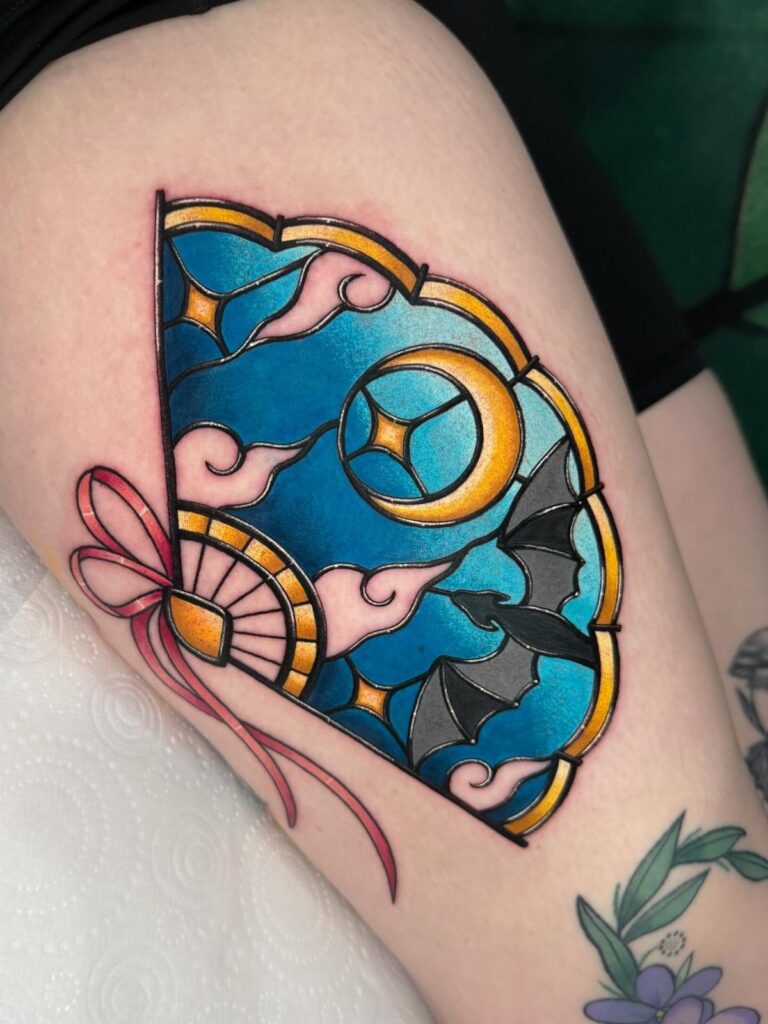 Stained glass fan tattoo on a thigh featuring a moon and bat silhouette.