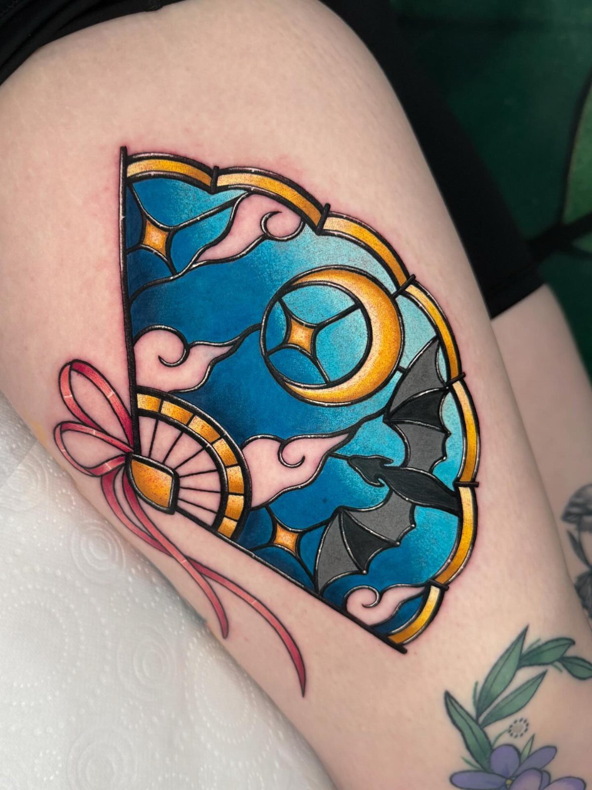 Stained glass fan tattoo on a thigh featuring a moon and bat silhouette.