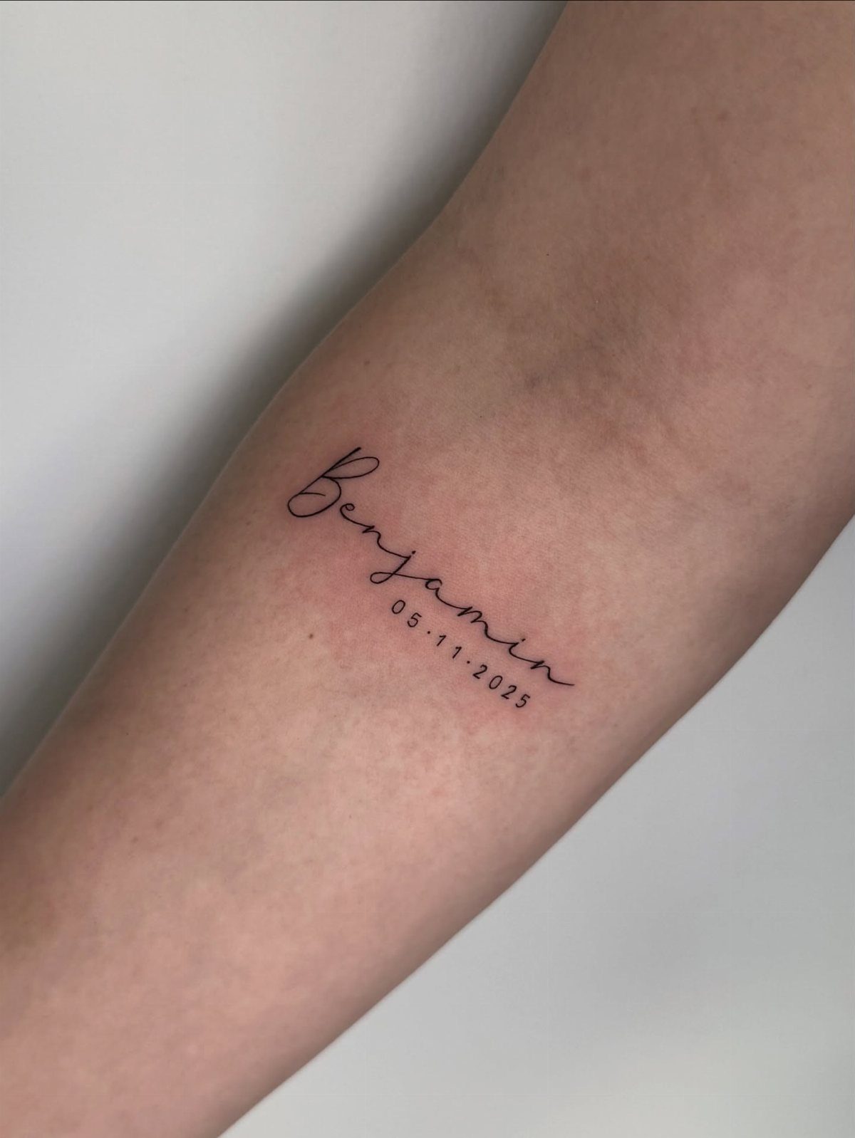 Cursive tattoo of the name Benjamin and a date on a forearm