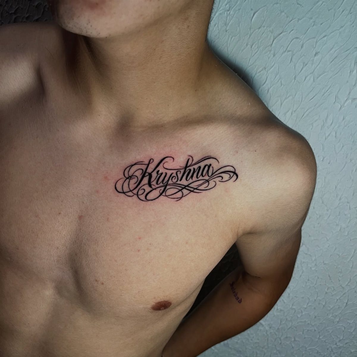 Black script name tattoo on a person's upper chest near the collarbone.