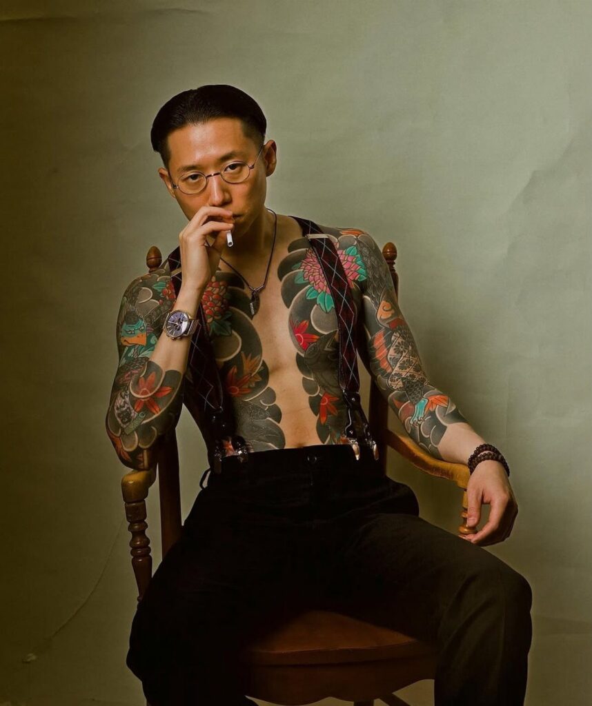 Man with traditional Japanese tattoos on chest and arms sitting in wooden chair