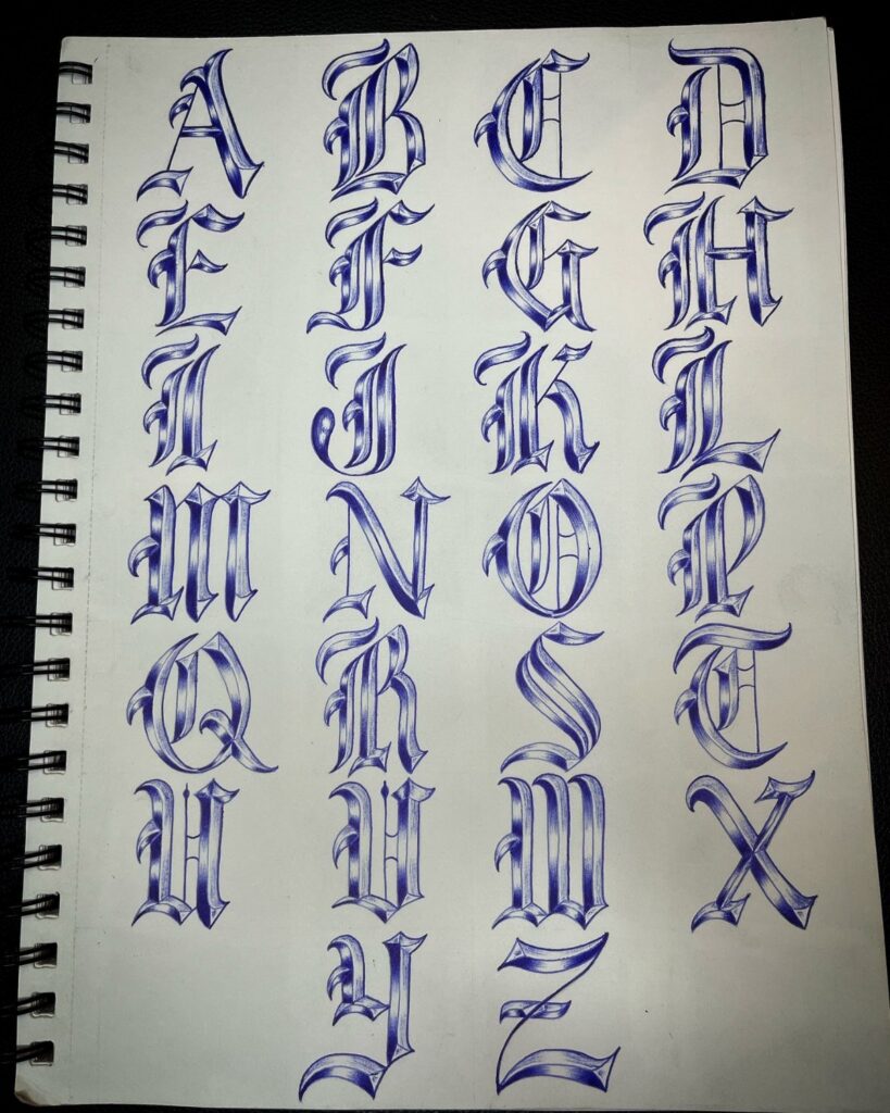 Hand-drawn blue and white traditional tattoo alphabet letters on a spiral sketchbook page