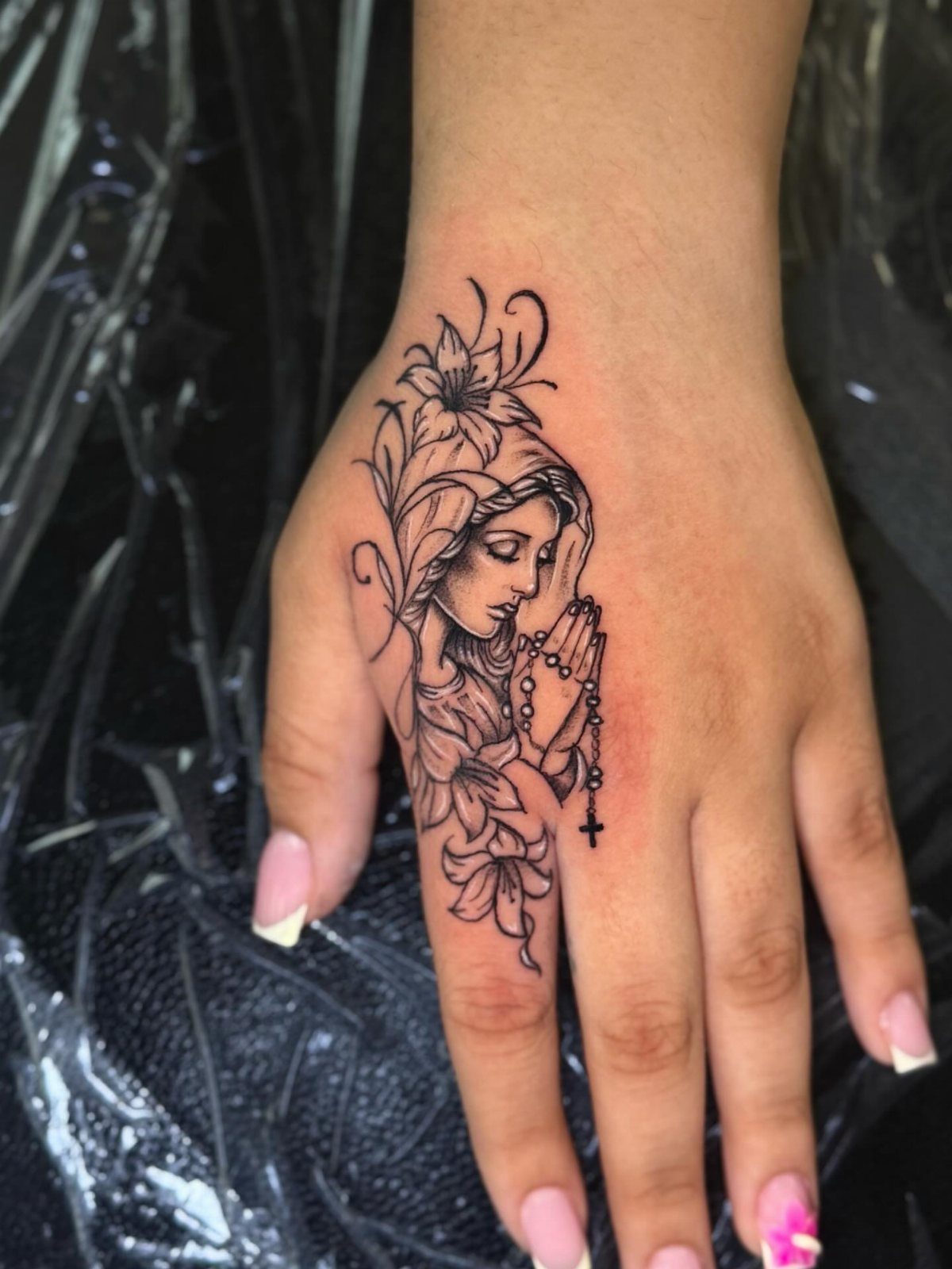 Virgin Mary tattoo with lilies and praying hands on the back of a hand