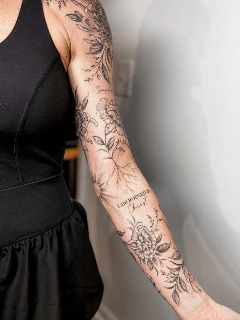 Woman's arm with floral tattoos and a religious script phrase on the forearm.